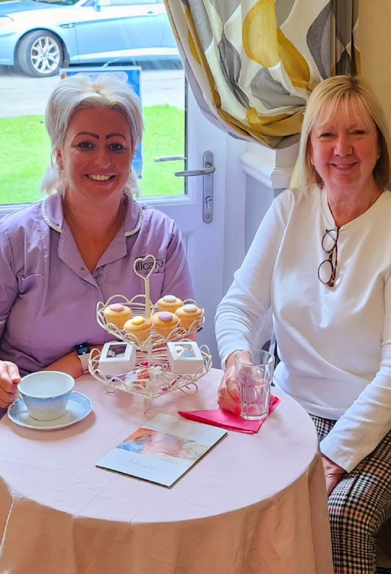 Two_women_smiling_sitting_at_a_small_table_drinking_tea_and_eating_scones