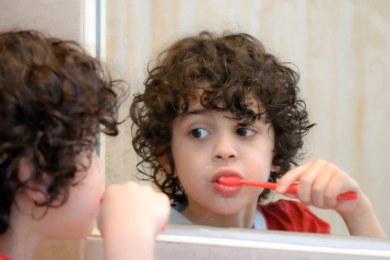 Young_boy_with_dark_curly_hair_looking_in_the_mirror_brushing_his_teeth_with_an_orange_toothbrush